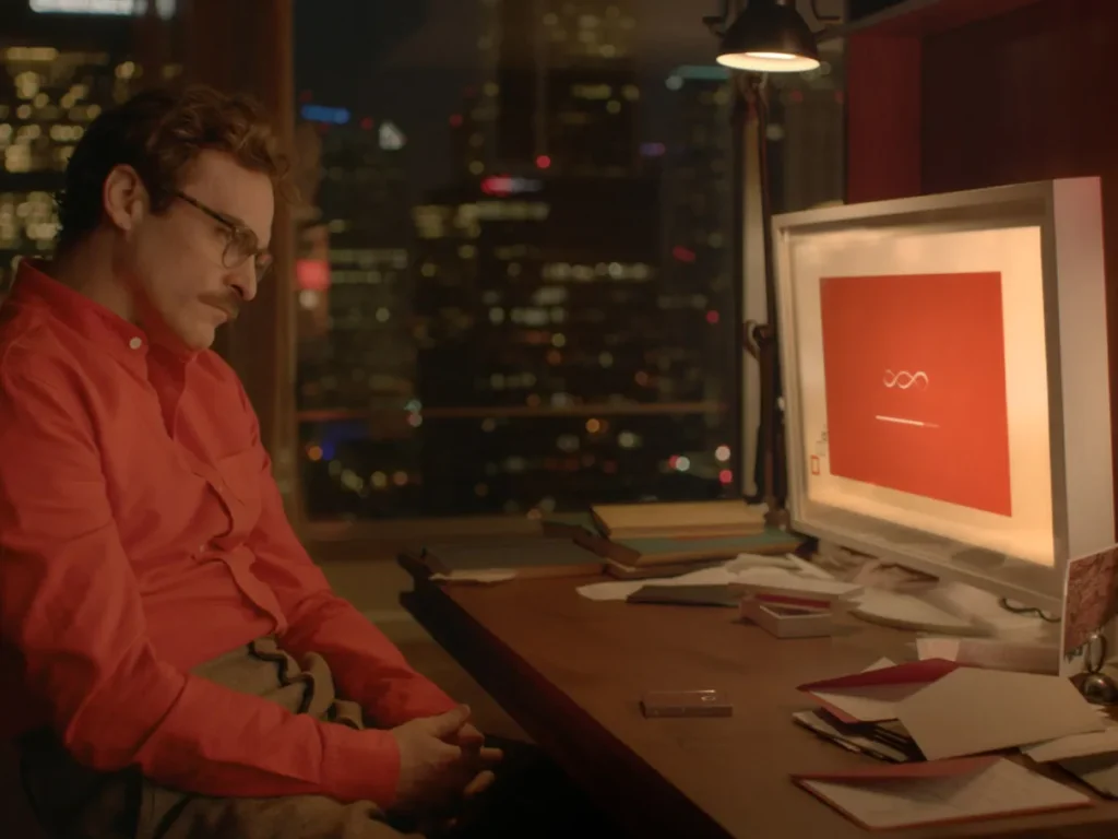 Homem de camisa vermelha sentado em frente a um monitor à noite, com vista da cidade ao fundo