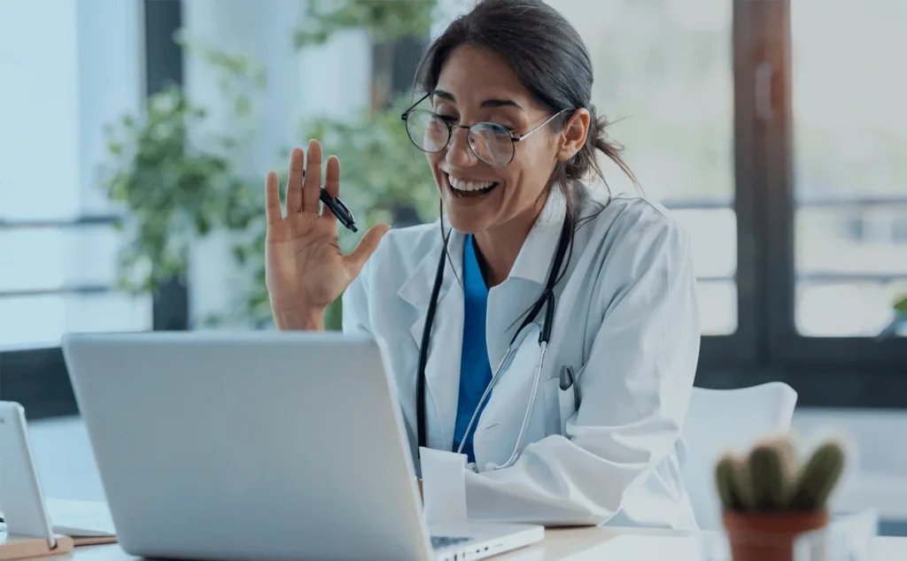 Médica de jaleco branco acena sorrindo durante videochamada no notebook