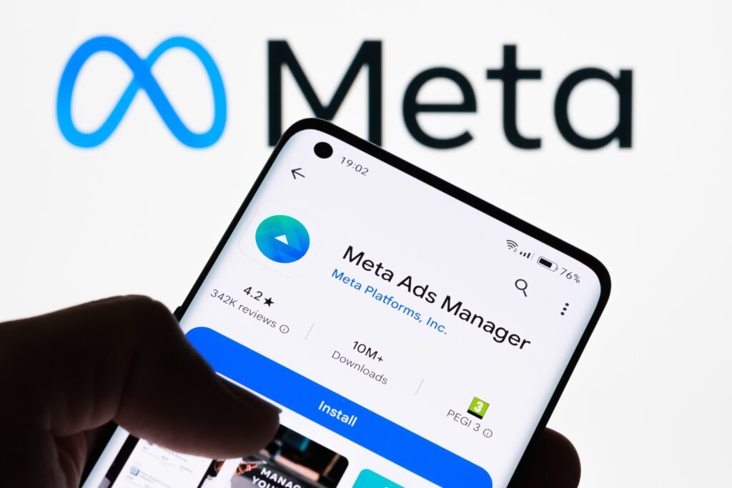 Mão segura celular exibindo o aplicativo Meta Ads Manager, com o logo da Meta ao fundo