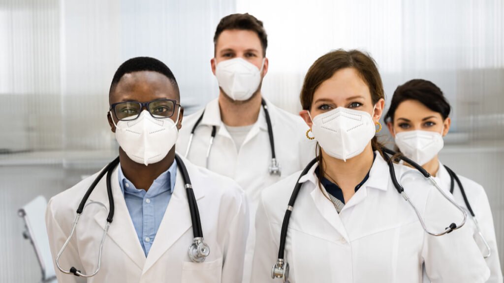 Quatro médicos de jaleco branco e máscara N95 posam lado a lado em consultório