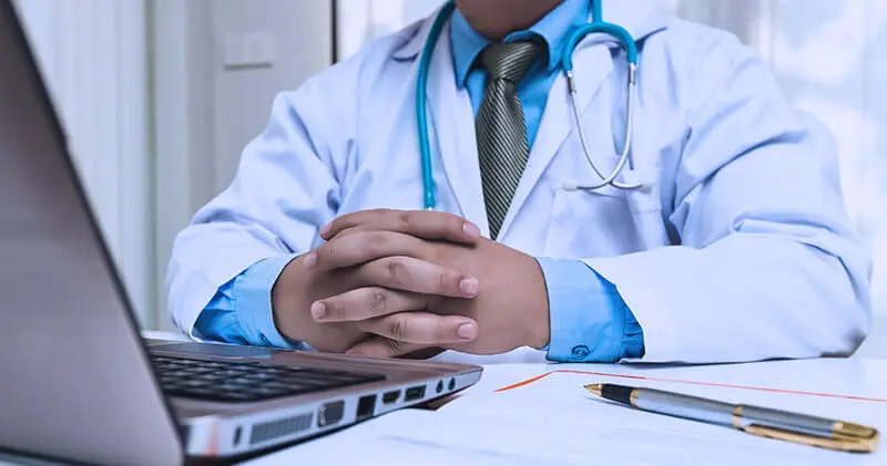 Médico de jaleco branco com as mãos entrelaçadas sobre a mesa, ao lado de notebook e caneta