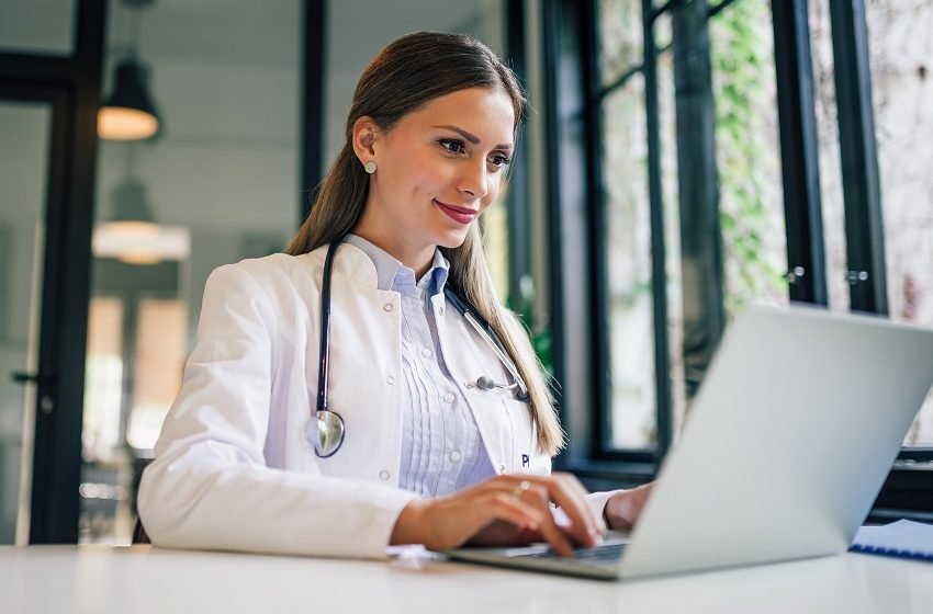 Médica sorridente digita no notebook em ambiente com plantas ao fundo