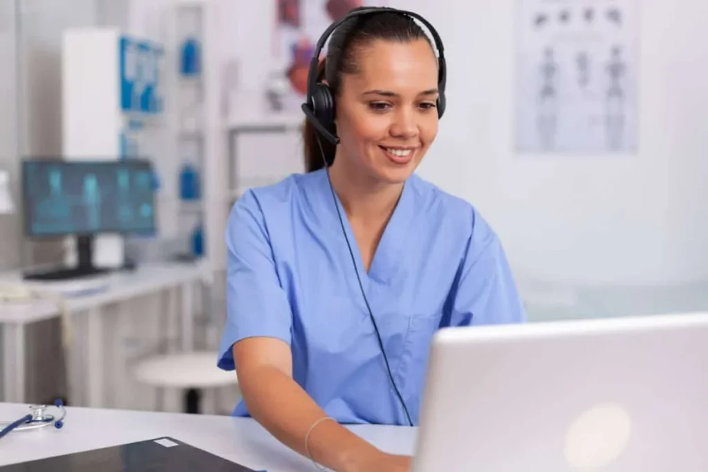 Enfermeira de scrubs azul com headset sorri em frente ao notebook em consultório