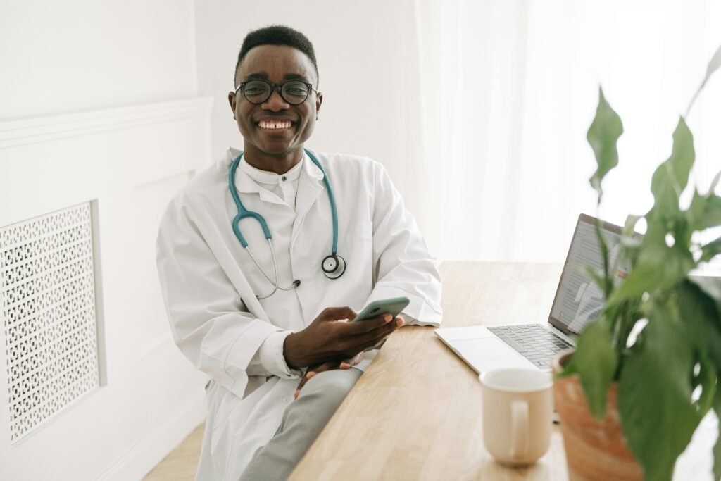 Médico sorridente de jaleco branco mexe no celular apoiado à mesa com notebook e planta