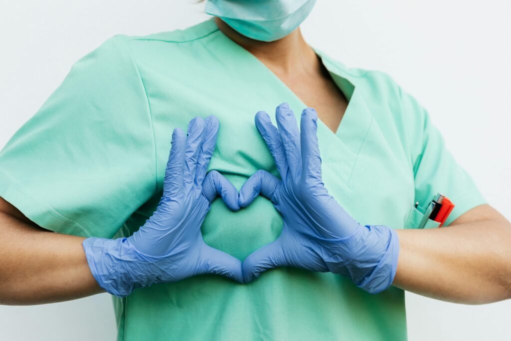 Profissional de scrubs verde-água faz um coração com as mãos enluvadas sobre o peito
