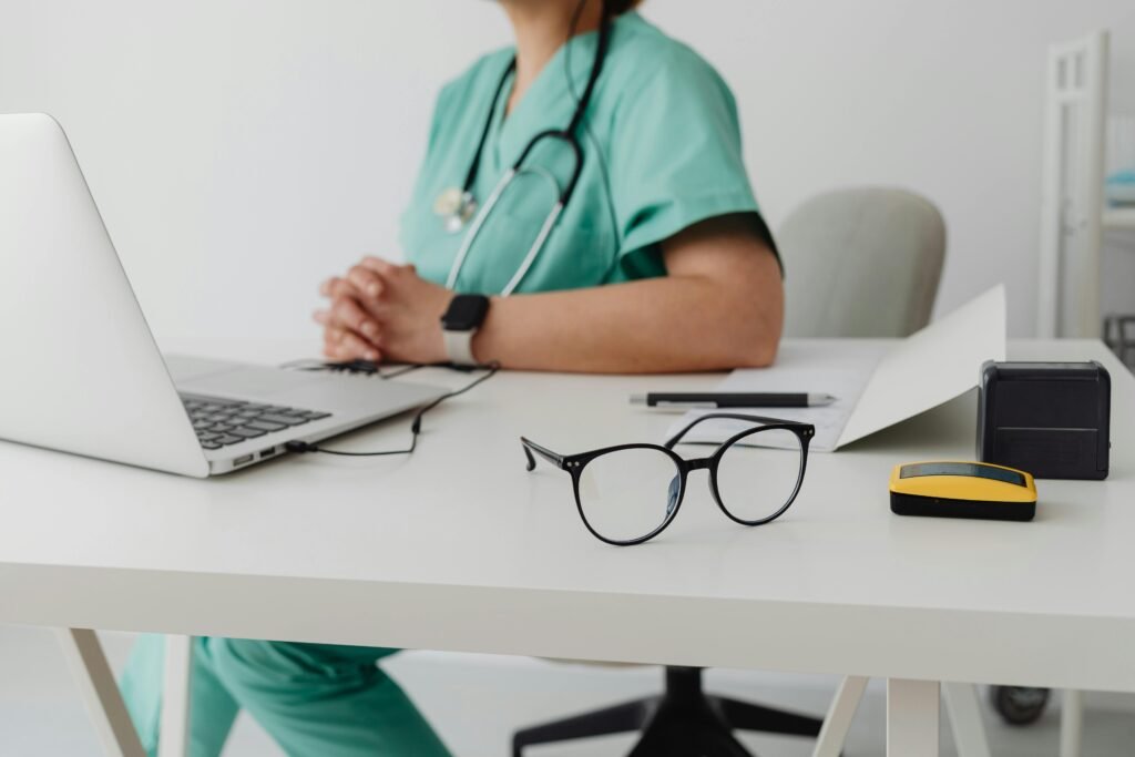 Enfermeira de scrubs verde-água ao computador, com óculos sobre a mesa em primeiro plano