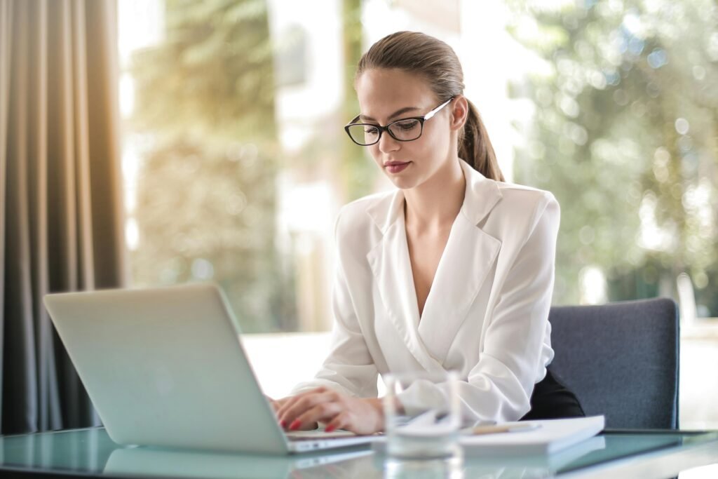 Mulher de blazer branco e óculos digita no notebook em mesa de vidro