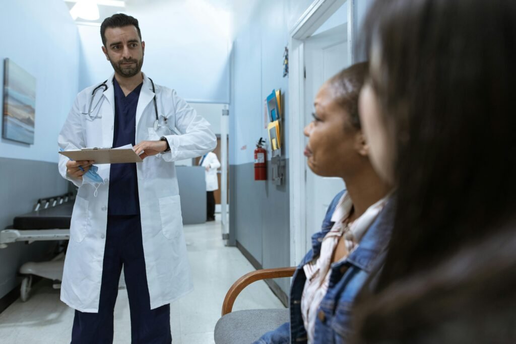 Médico com prancheta conversa com duas pacientes sentadas em sala de espera de clínica