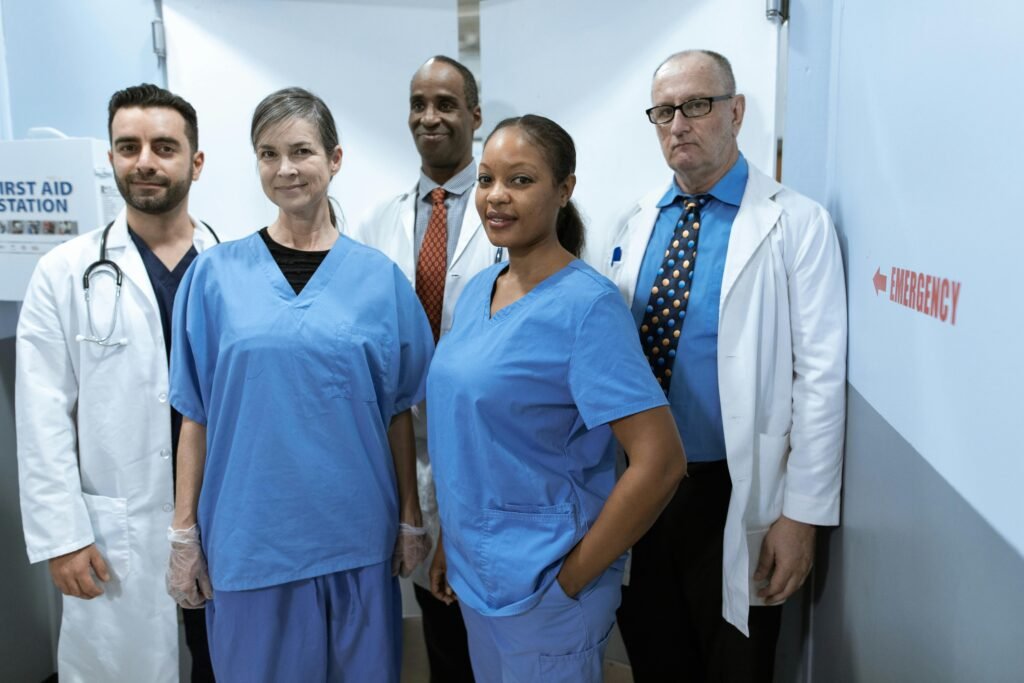 Cinco profissionais de saúde de jaleco e scrubs azul posam de braços cruzados em corredor de hospital