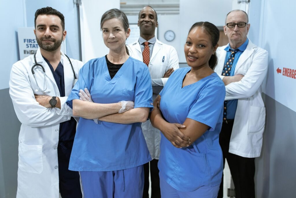 Cinco profissionais de saúde de jaleco e scrubs azul posam de braços cruzados em corredor de hospital