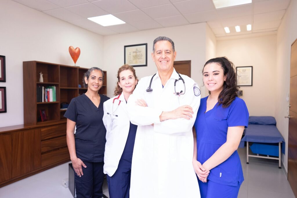 Equipe de quatro profissionais de saúde de jaleco e scrubs posa sorrindo em consultório