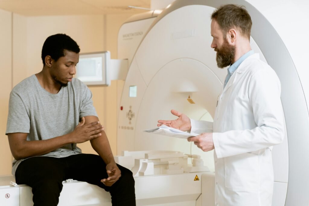 Médico com prancheta conversa com paciente sentado ao lado de aparelho de ressonância magnética