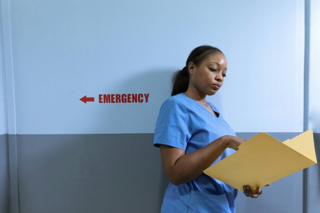 Enfermeira de scrubs azul lê uma pasta amarela ao lado de placa indicando 'Emergency'
