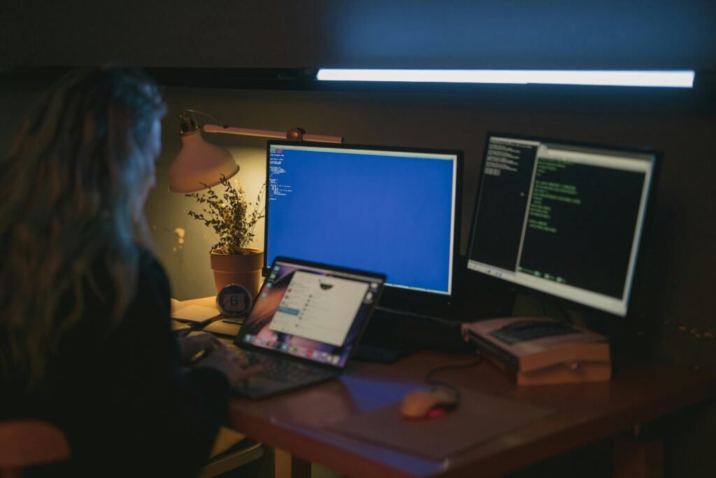Pessoa trabalha à noite em mesa com notebook e dois monitores exibindo código de programação