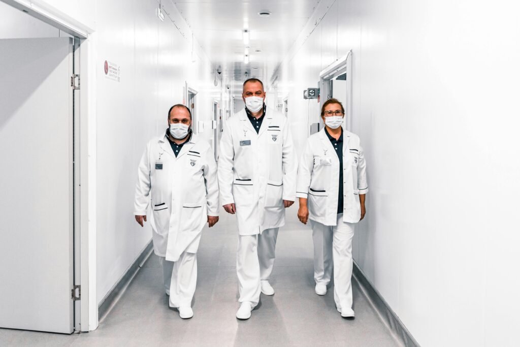 Três profissionais de saúde de máscara e jaleco branco caminham por corredor de hospital