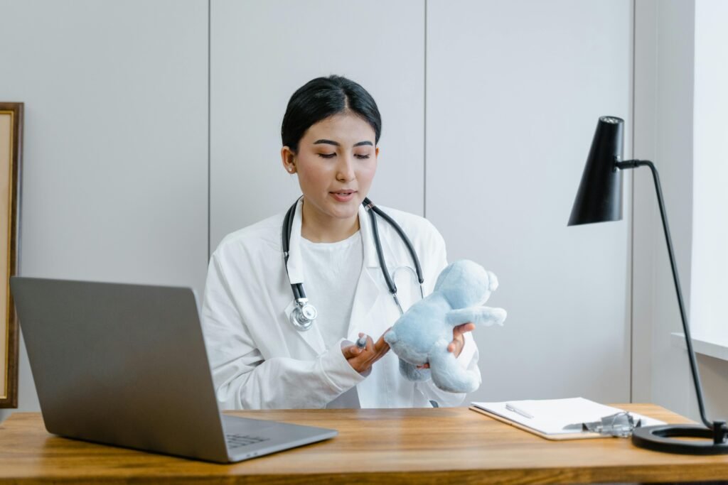 Médica de jaleco branco segura um ursinho de pelúcia azul enquanto conversa em videochamada no notebook