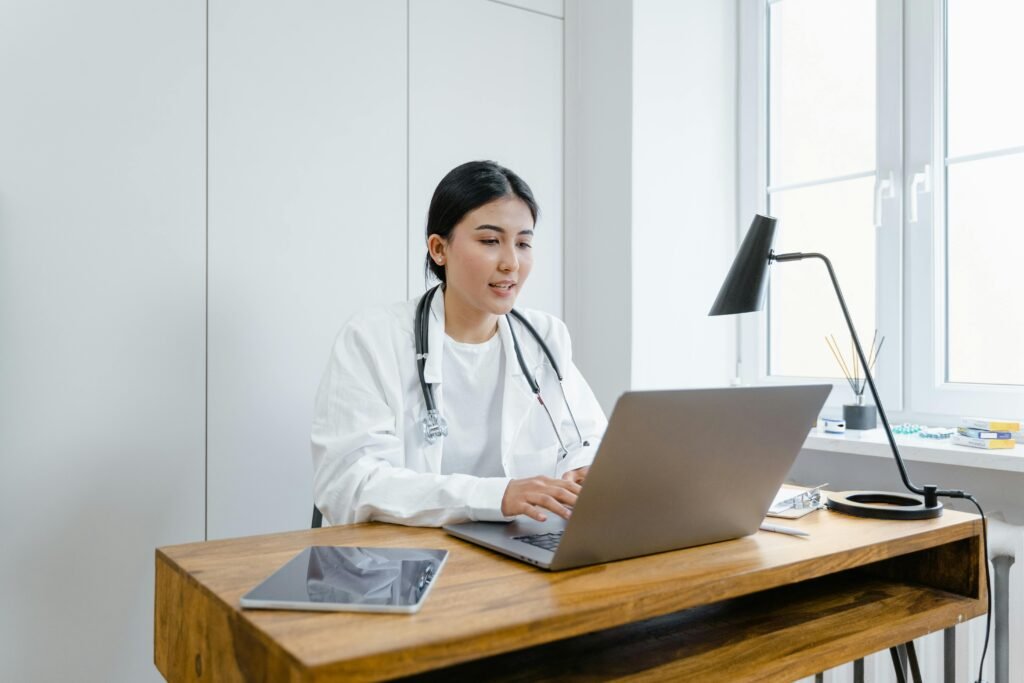 Médica de jaleco branco digita no notebook em mesa de madeira