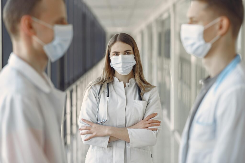 Médica de máscara e estetoscópio, braços cruzados, entre dois colegas em corredor de hospital