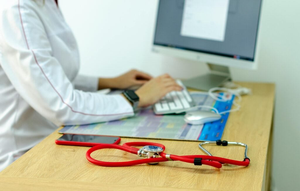 Estetoscópios vermelho e cinza sobre mesa de madeira em consultório, com médica digitando ao computador ao fundo