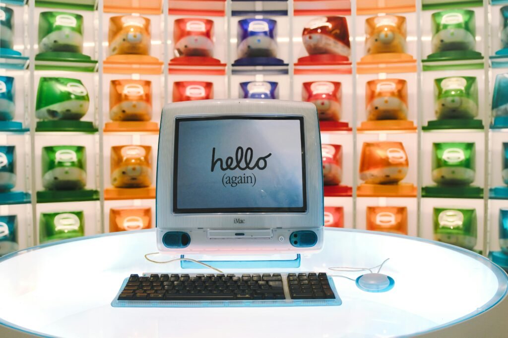 iMac G3 vintage exibindo a tela 'hello (again)' em exposição de computadores coloridos