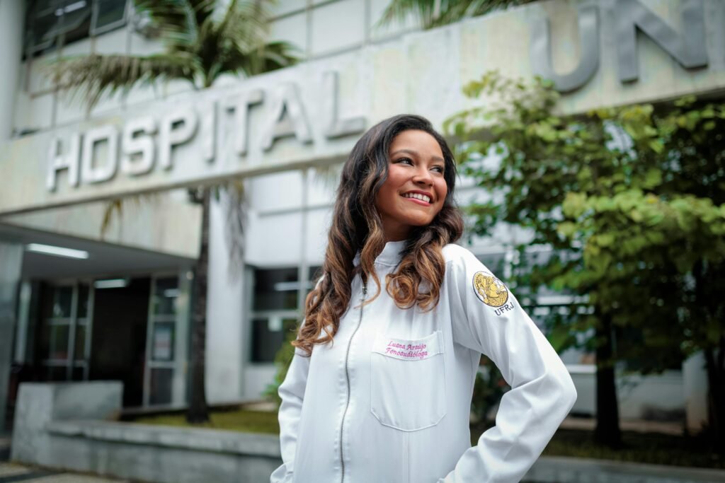 Fonoaudióloga sorridente de jaleco branco em frente à fachada de um hospital universitário