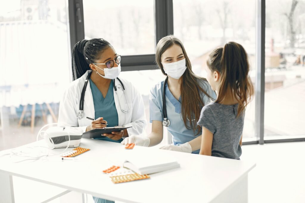 Duas médicas de máscara conversam com uma menina em consulta pediátrica, com remédios sobre a mesa