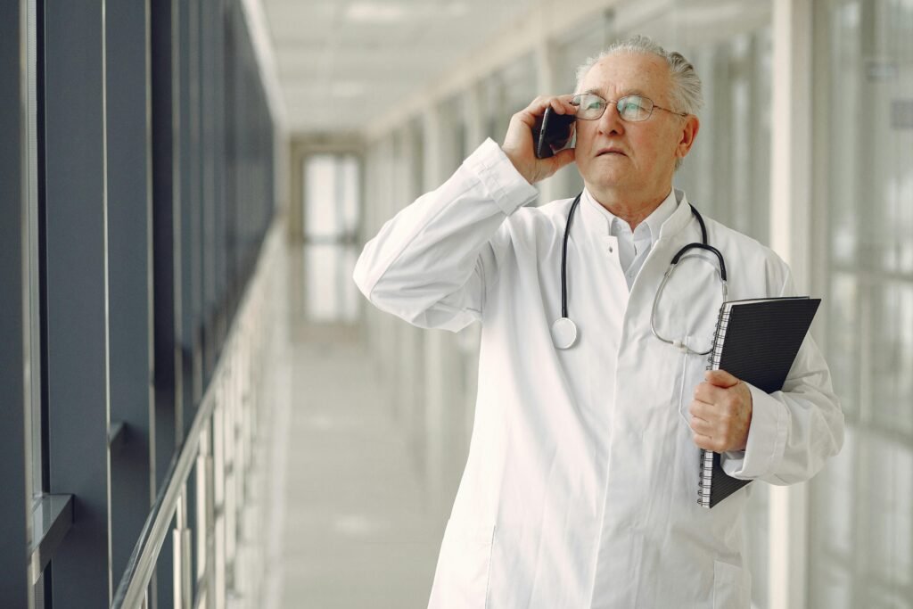 Médico idoso fala ao telefone enquanto caminha pelo corredor do hospital com um caderno em mãos