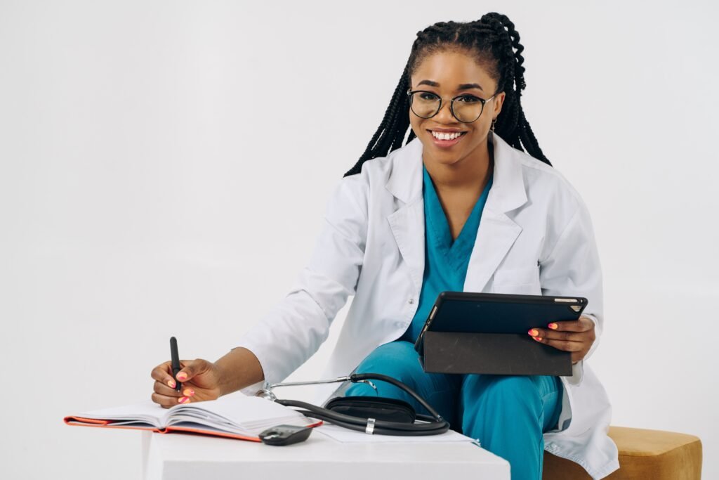 Médica sorridente segurando tablet e anotando em caderno sobre a mesa, com estetoscópio ao lado