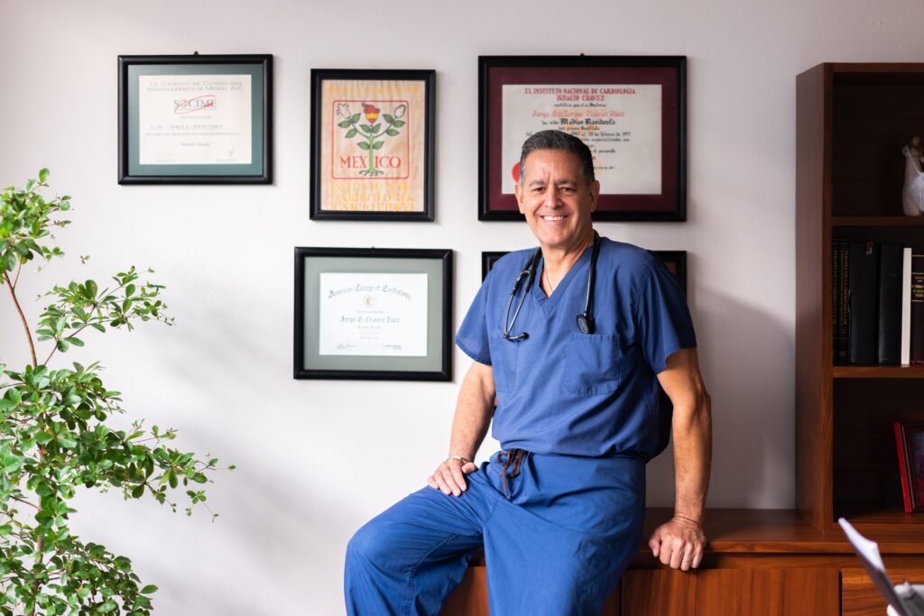 Médico cardiologista de scrubs azul sorrindo em seu consultório, com diplomas emoldurados na parede