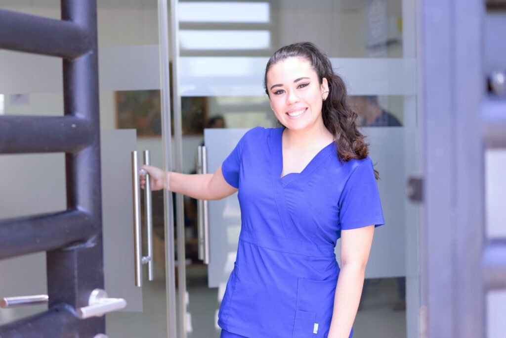 Profissional de saúde de scrubs azul sorrindo na entrada de uma clínica, segurando a porta para receber pacientes