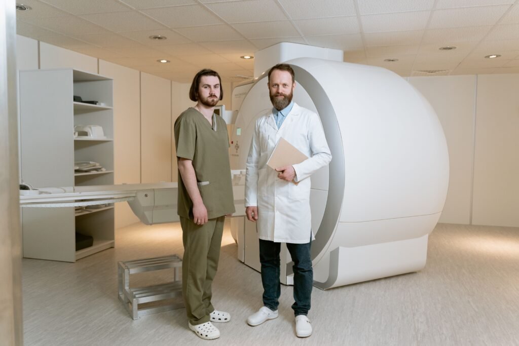 Médico de jaleco branco e técnico de scrubs verdes ao lado de um aparelho de ressonância magnética em sala de exame